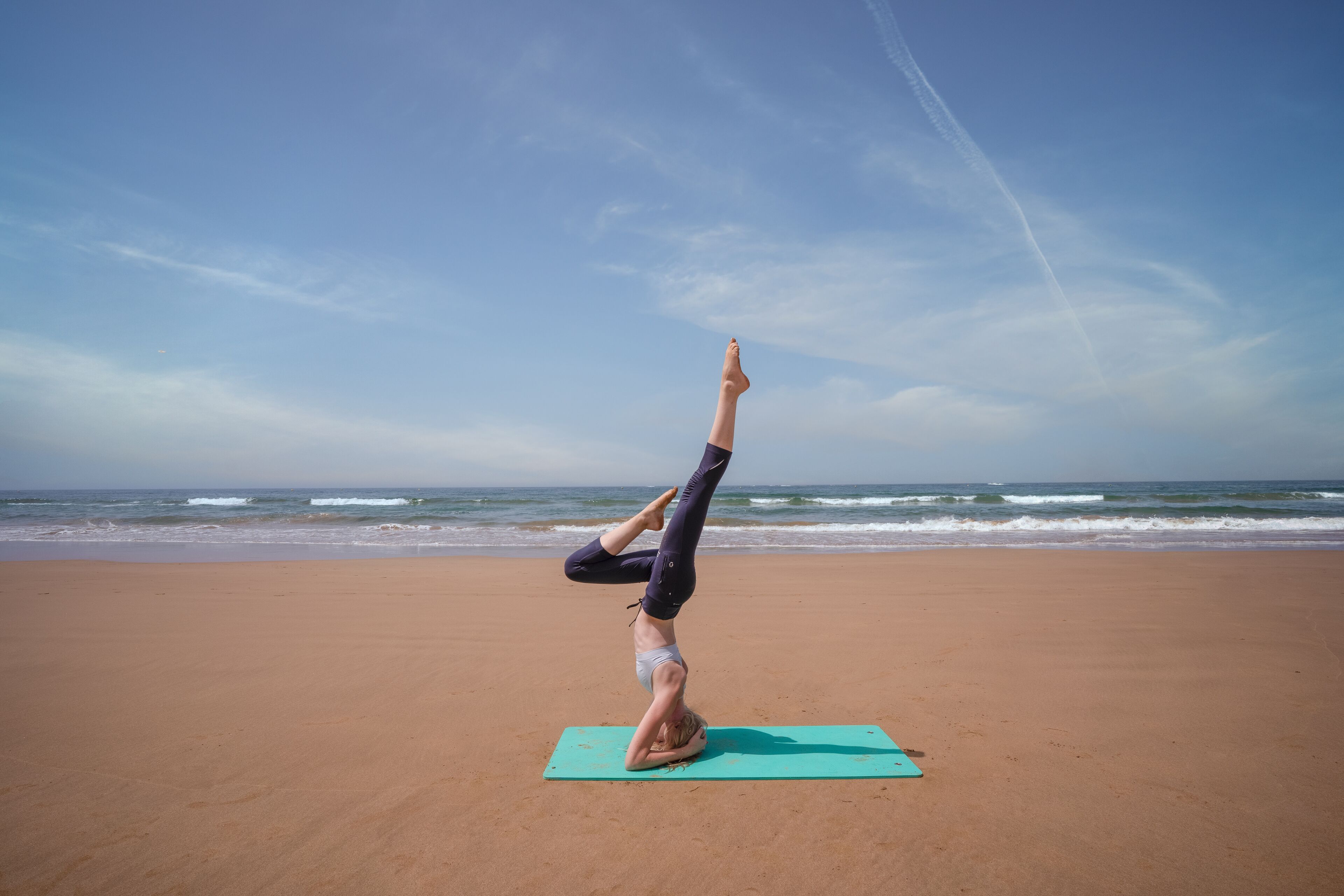 A person performing a handstand on a yoga mat on a beach with the ocean in the background.