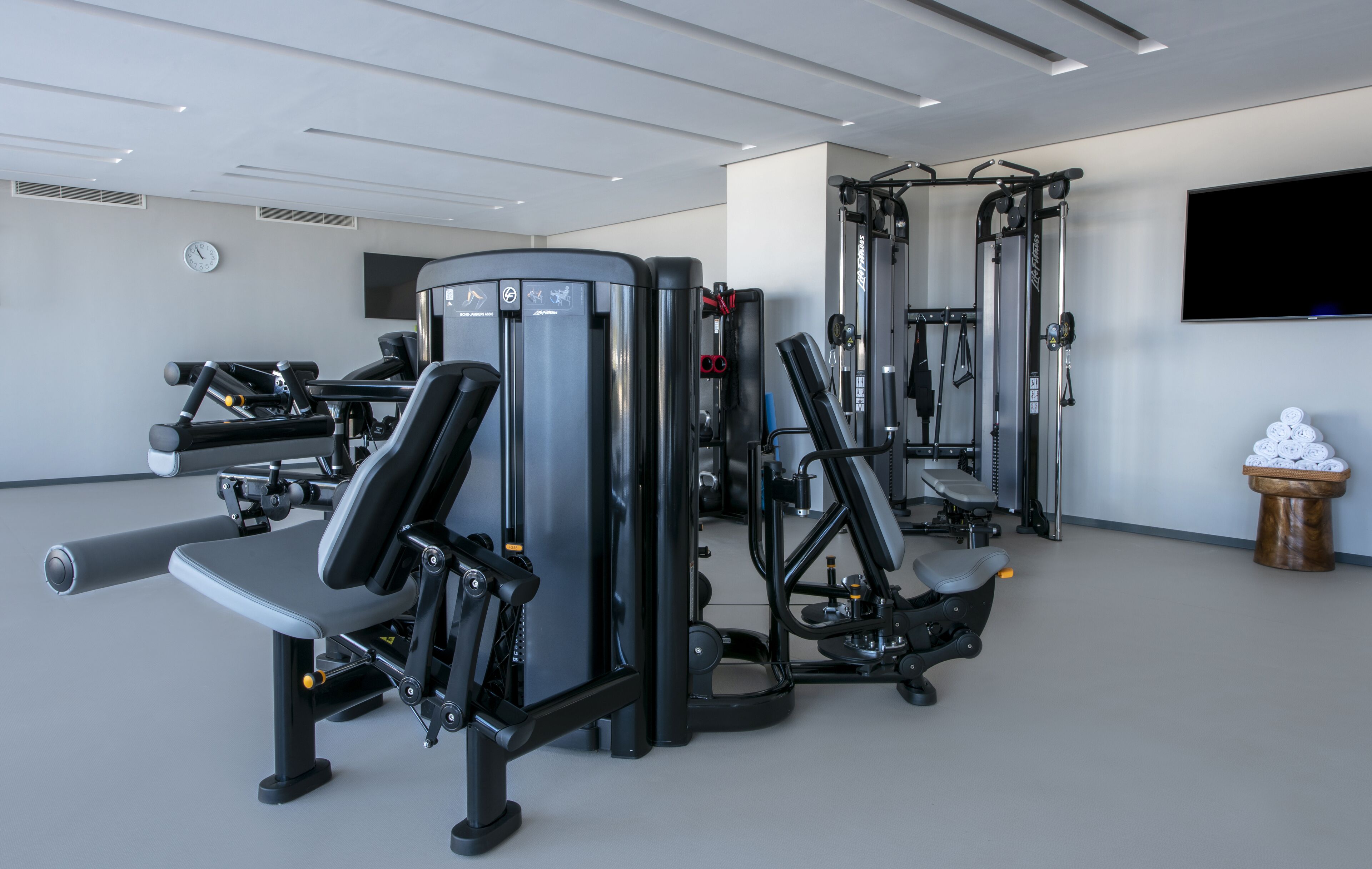 Et fitnessrum med forskellige træningsmaskiner, et fjernsyn ophængt på væggen og et ur.