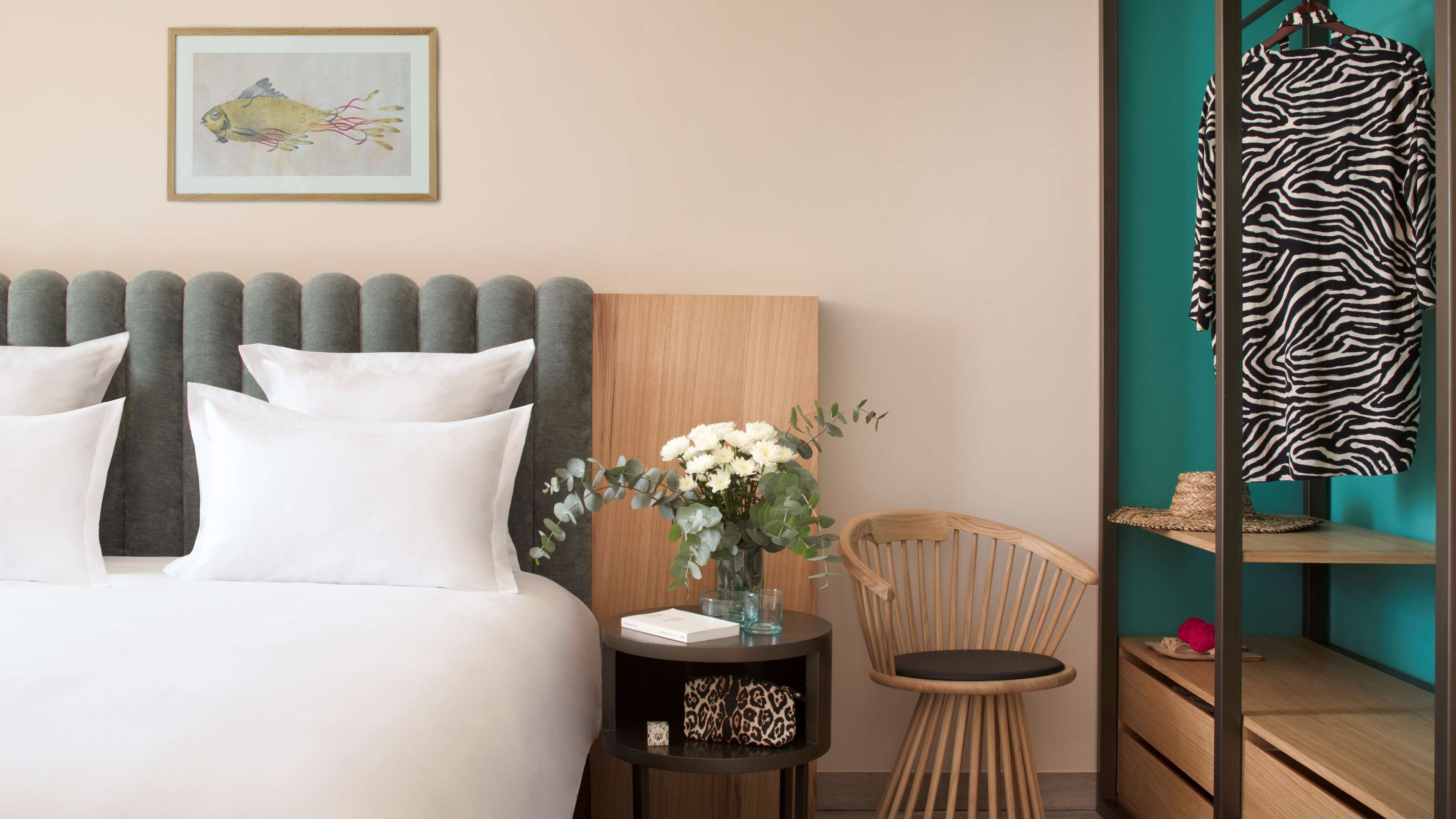 A neatly arranged hotel room with a bed, a side table with a vase of flowers, a chair, and a zebra-patterned garment hanging on a rack.