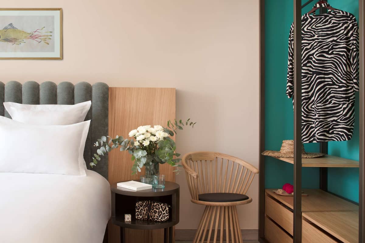 A neatly arranged hotel room with a bed, a side table with a vase of flowers, a chair, and a zebra-patterned garment hanging on a rack.