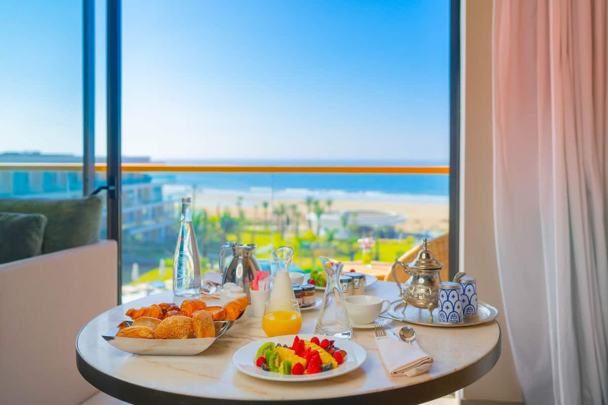 A breakfast table with a variety of food items and drinks, a pitcher, and a teapot, set against a view of a beach and ocean.