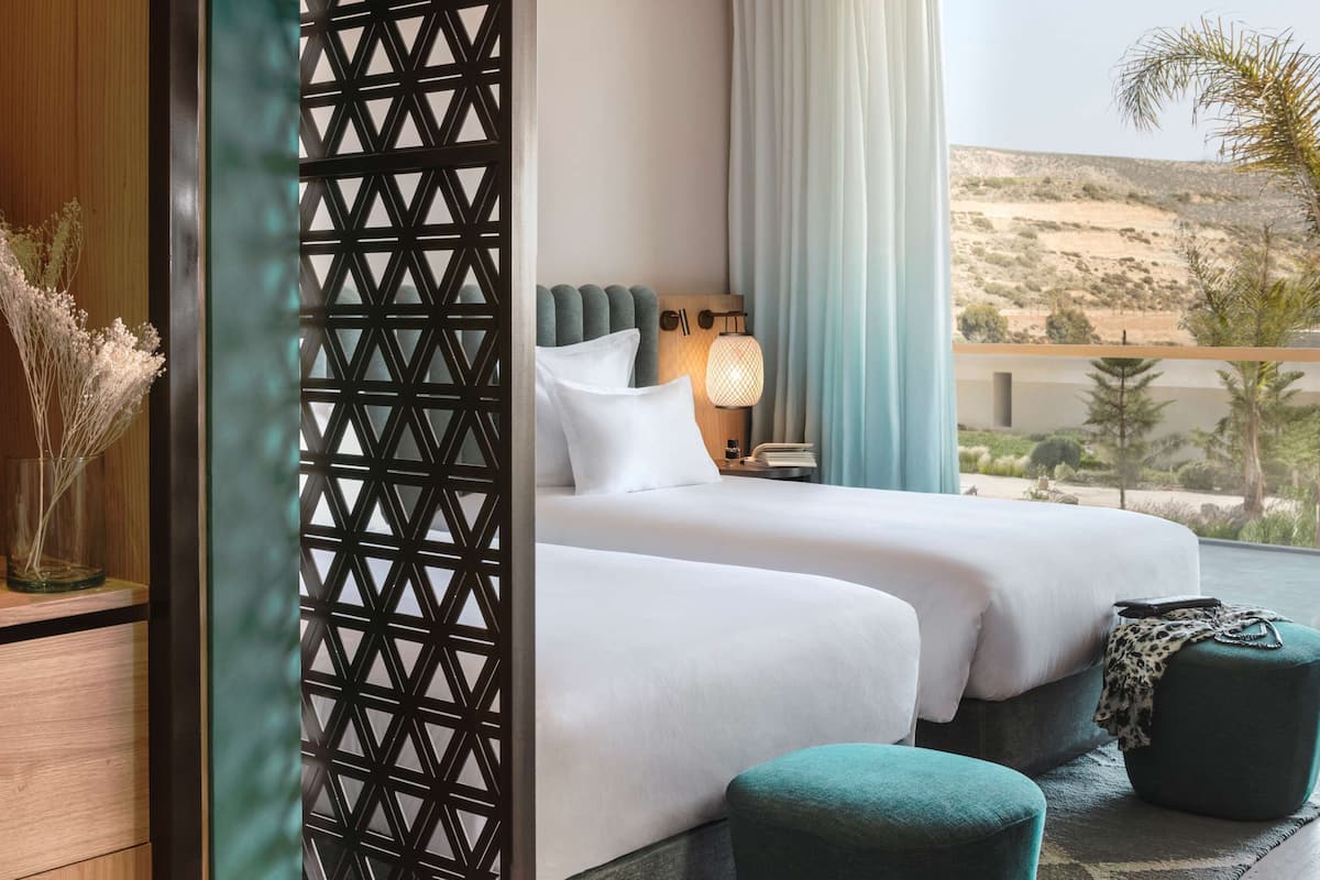 A bedroom with two beds, a view of the desert, a wooden shelf with a vase, and a decorative screen.