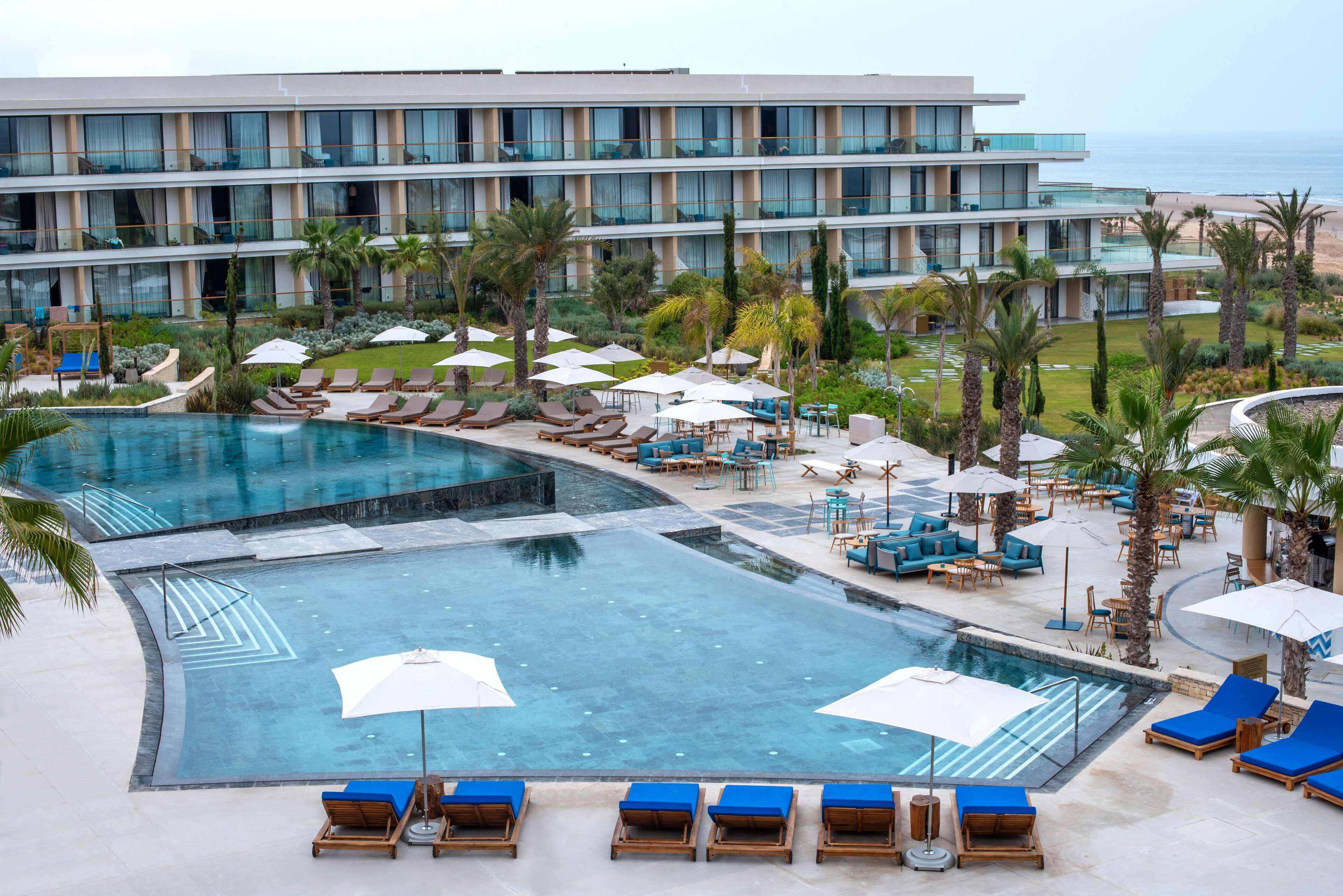 A hotel with a large swimming pool, sun loungers, umbrellas, and a view of the ocean.