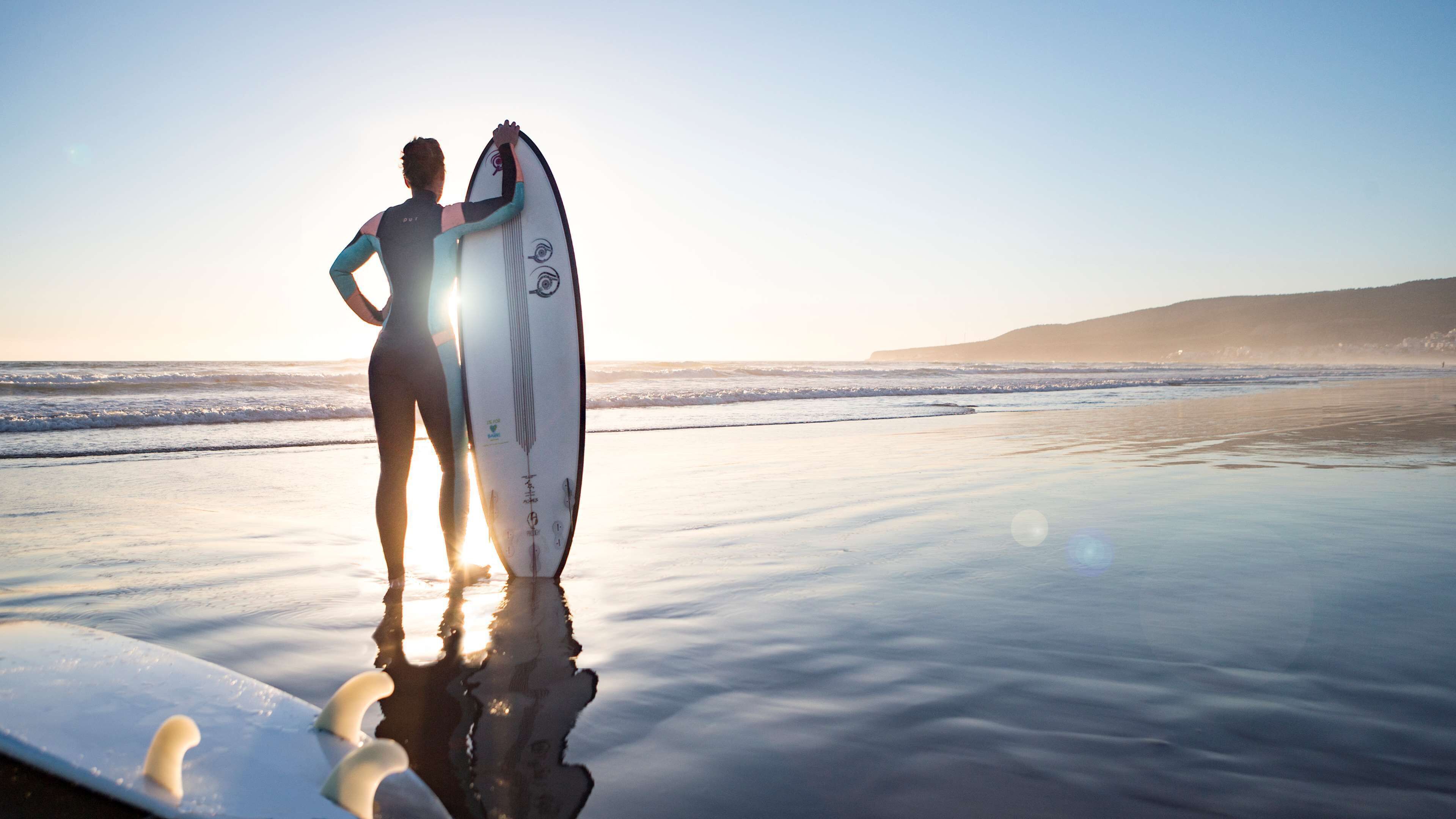 Una persona in piedi sulla spiaggia con una tavola da surf, che si specchia nell'acqua.
