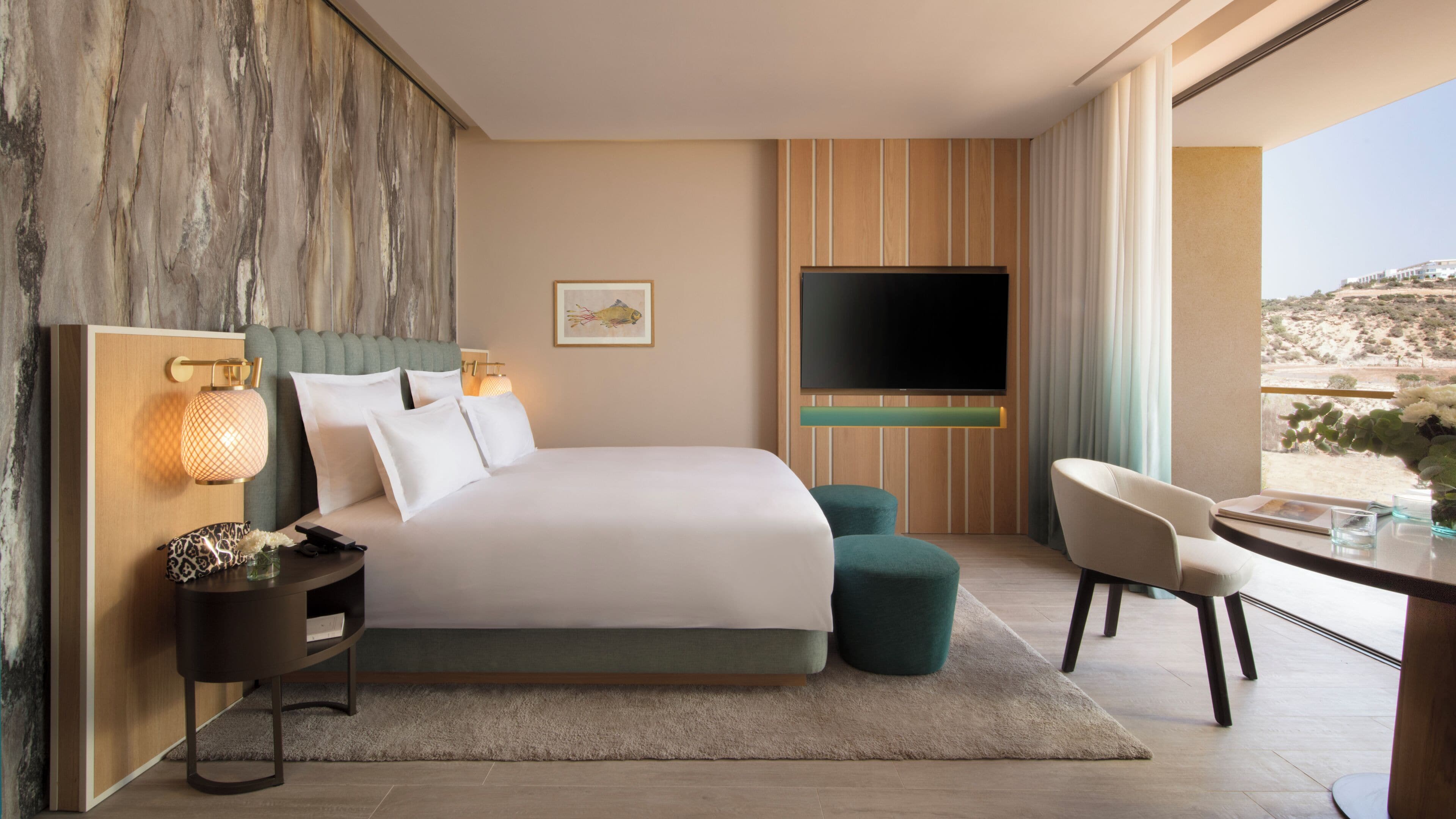 A modern hotel room with a large bed, a flat-screen TV, a desk with a chair, and a view of the desert.