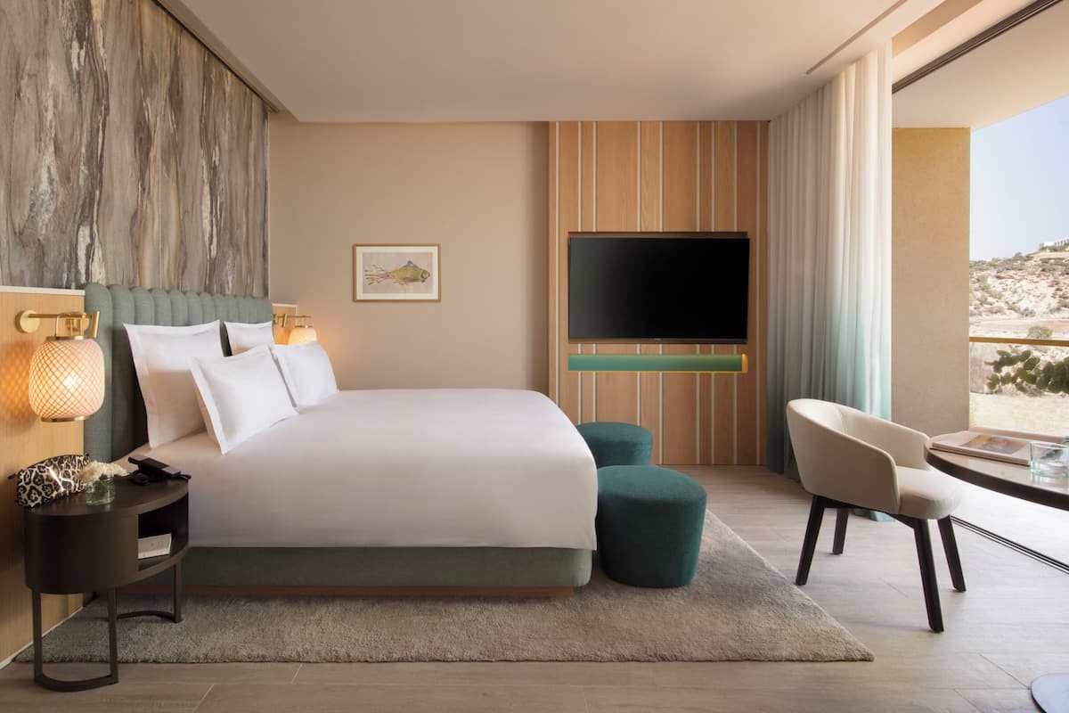 A modern hotel room with a large bed, a flat-screen TV, a desk with a chair, and a view of the desert.