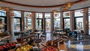 A spacious hotel lobby with multiple seating areas, large windows offering views of the outdoors, and a variety of fruits and pastries displayed on tables. - Hyatt Regency Taghazout  (Taghazout)