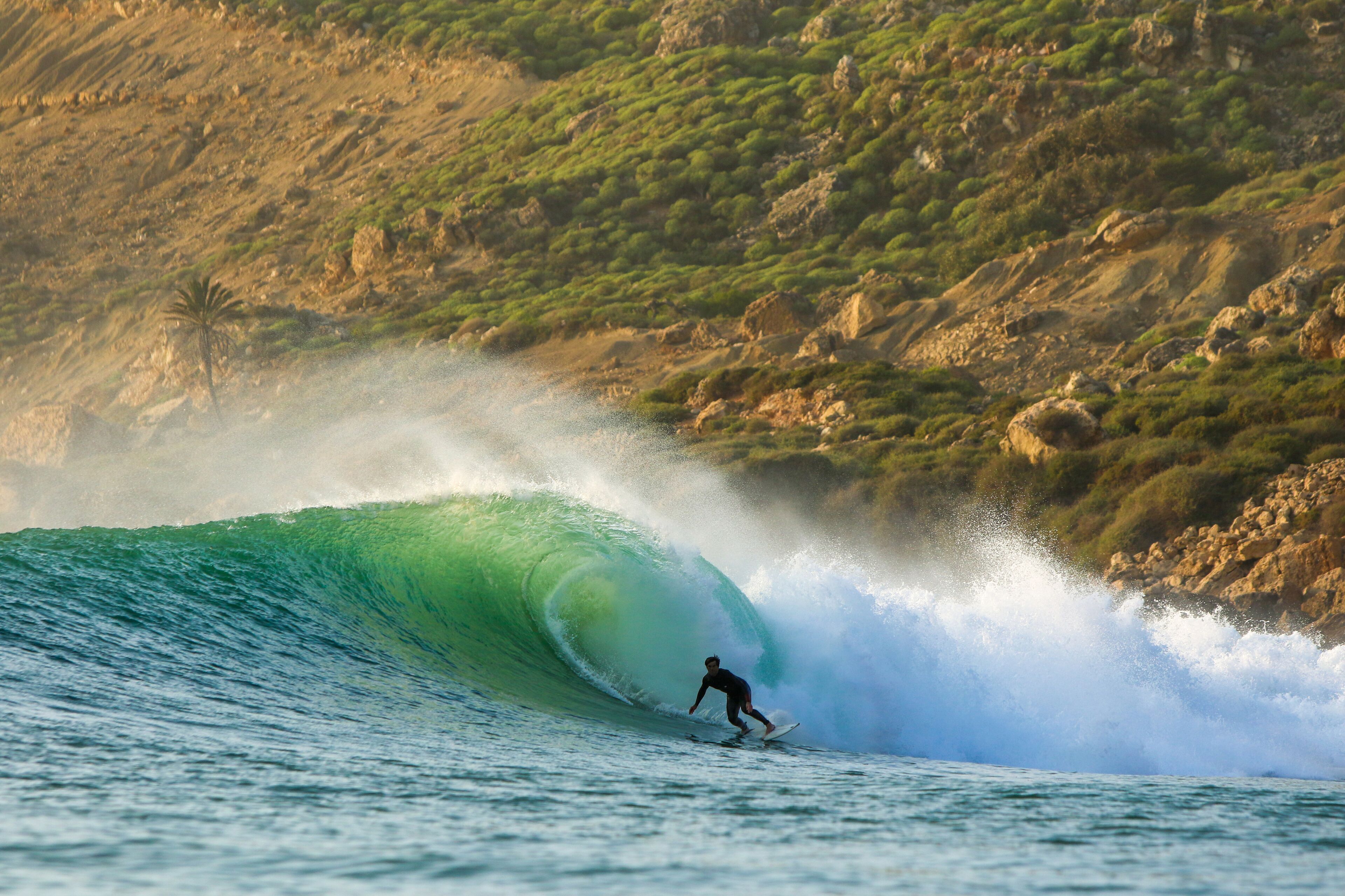 A surfer riding a large wave with a rocky hillside in the background.