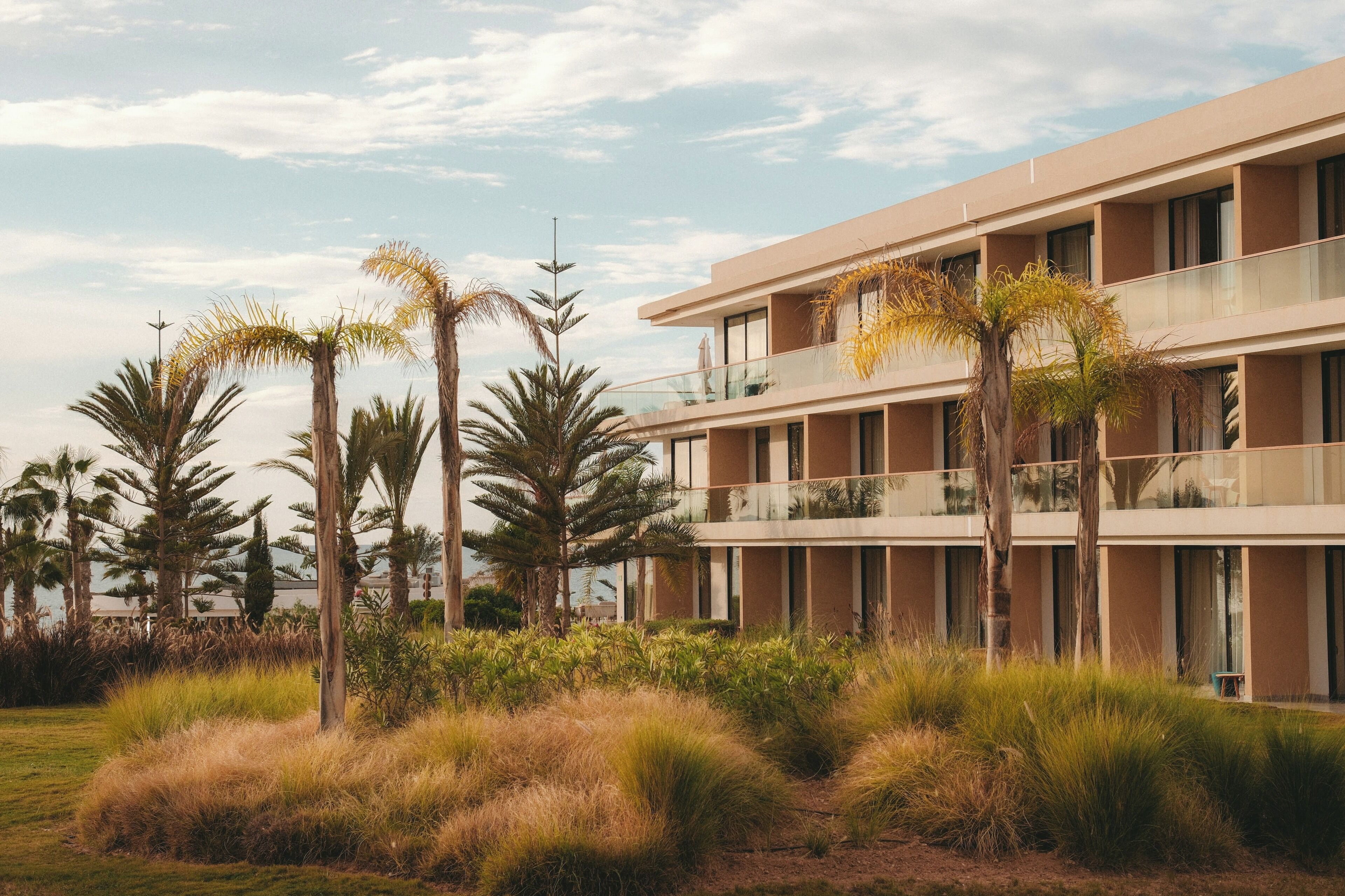 A modern hotel with multiple balconies, palm trees, and a grassy area.
