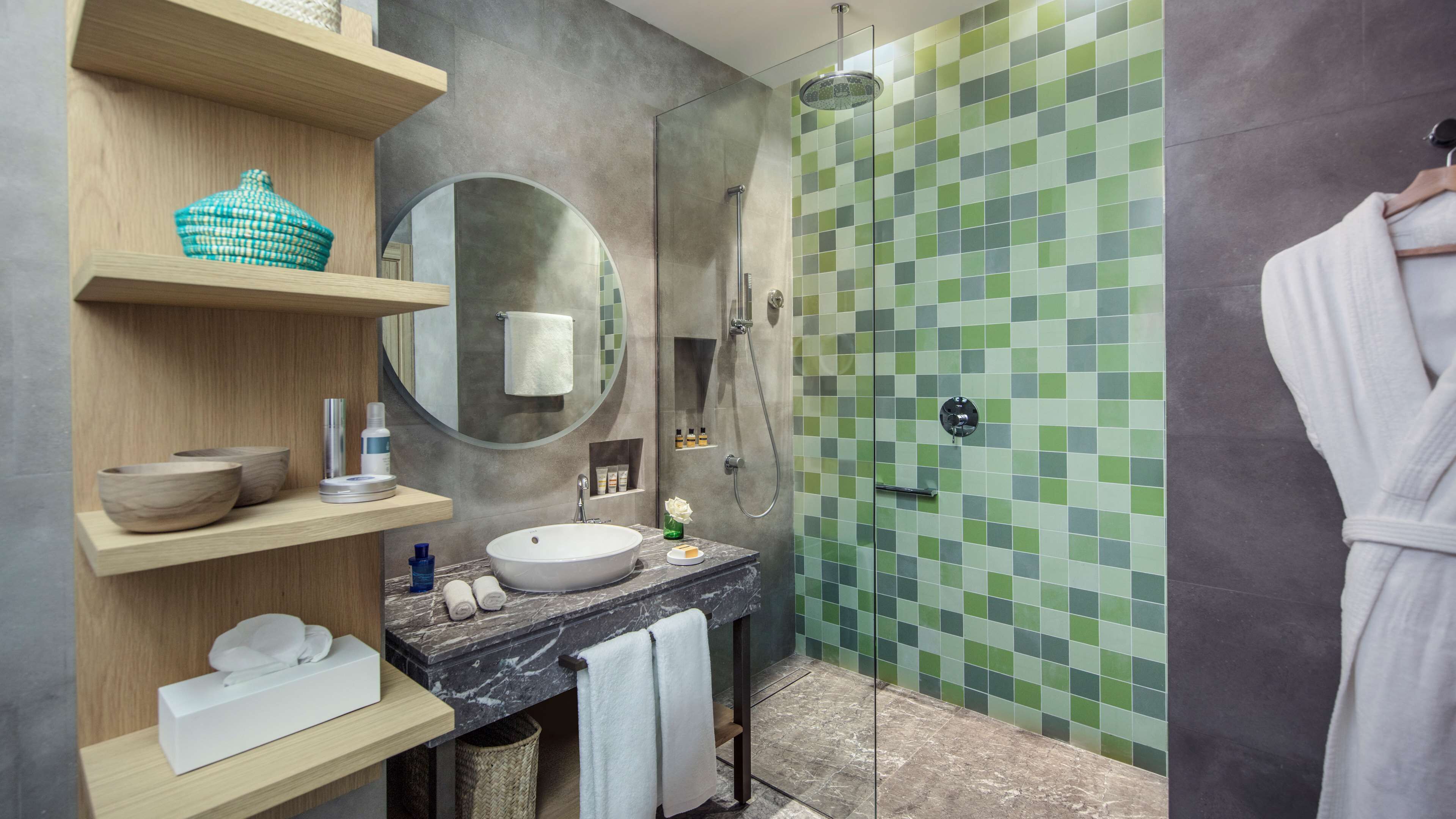 Un baño moderno con un lavabo de mármol, estanterías de madera, un espejo redondo y una ducha con azulejos de mosaico verde.