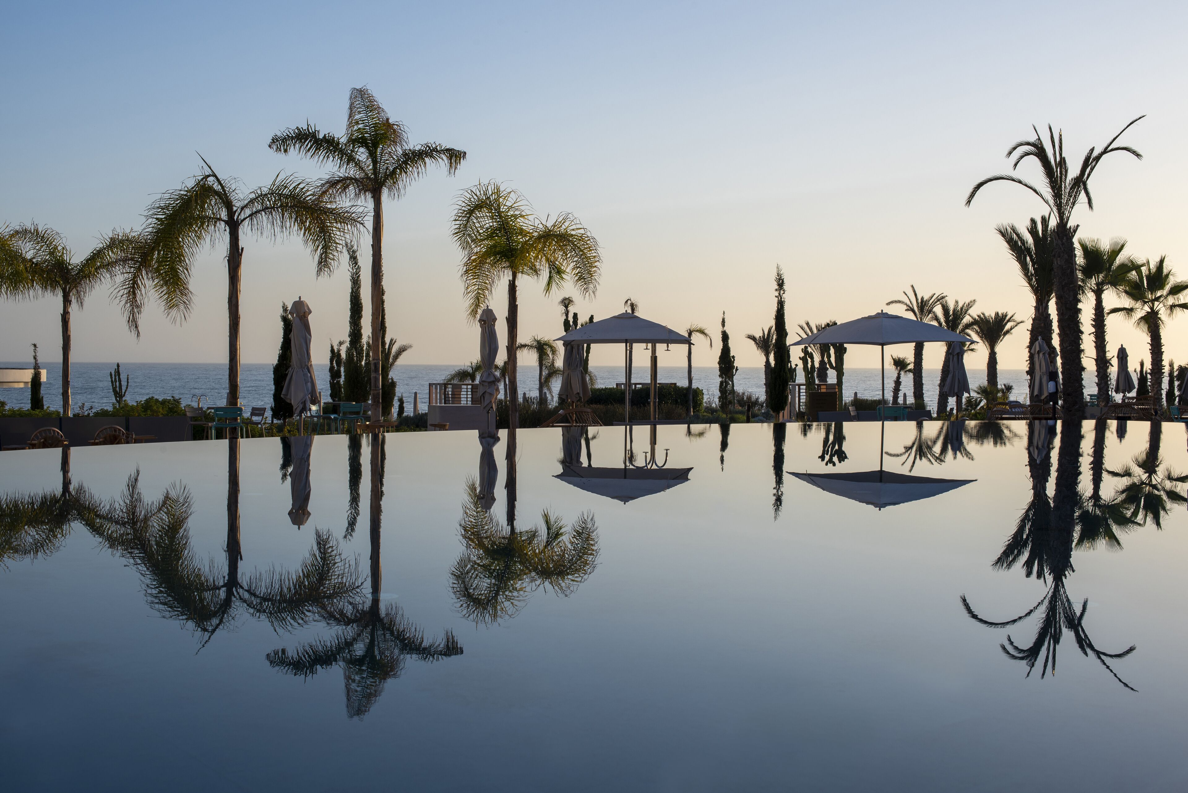 A serene pool area with palm trees and gazebos, reflecting the sky and surroundings.
