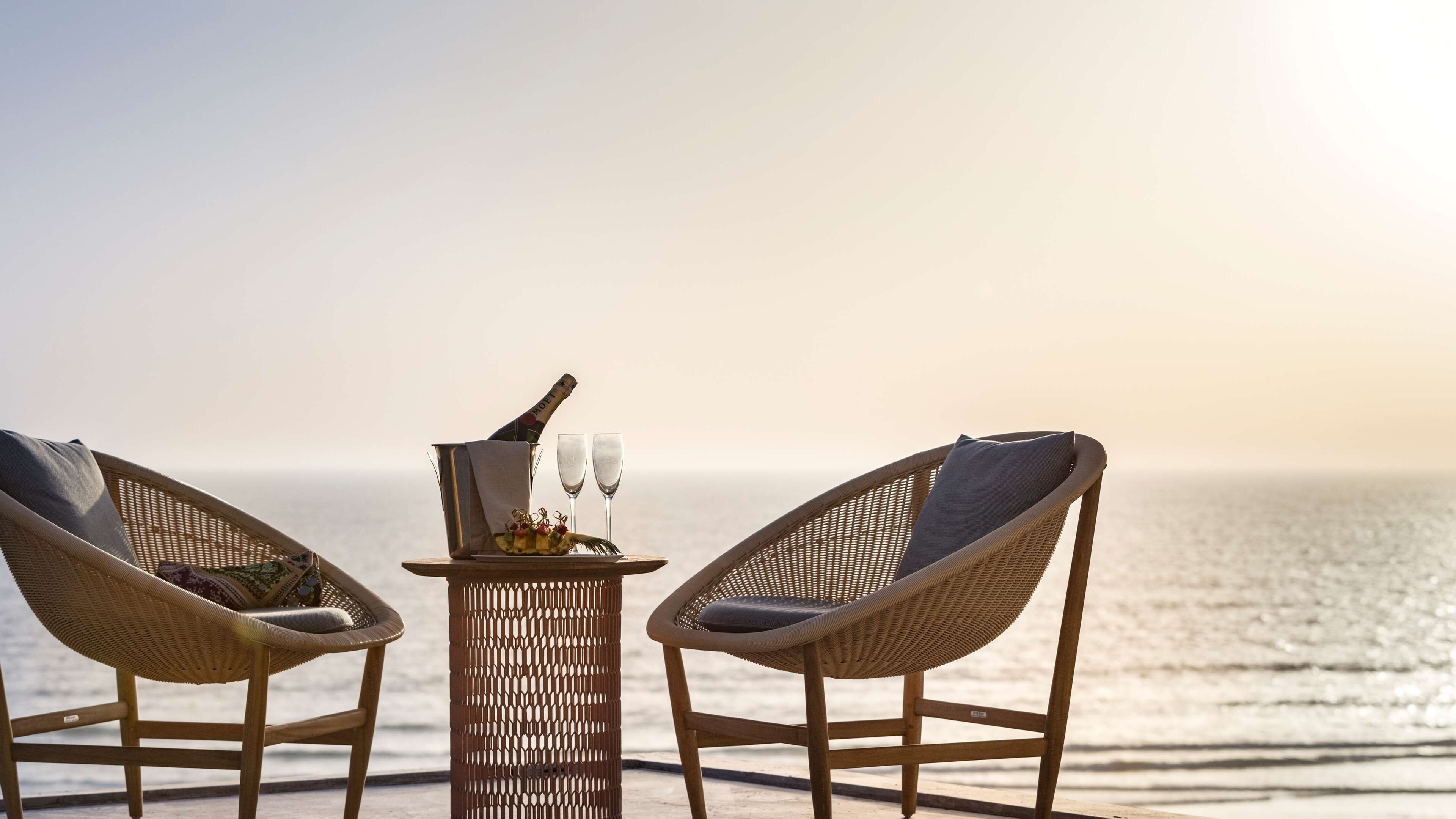 Two wicker chairs facing a table with a bottle of champagne and two glasses, set on a deck with a view of the ocean.