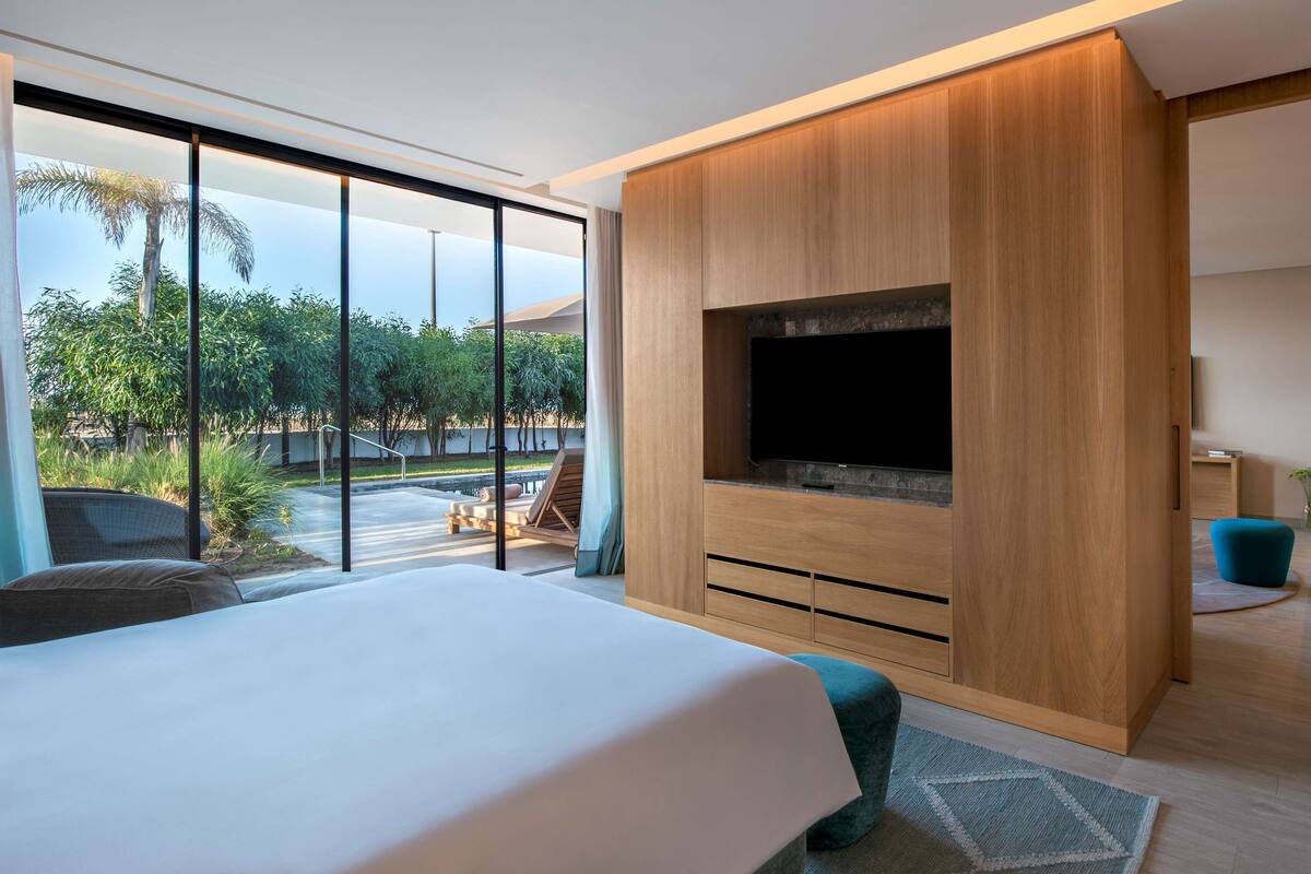 A modern bedroom with a large bed, a TV mounted on a wooden cabinet, and a view of a pool and greenery outside.