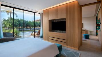 A modern bedroom with a large bed, a TV mounted on a wooden cabinet, and a view of a pool and greenery outside.