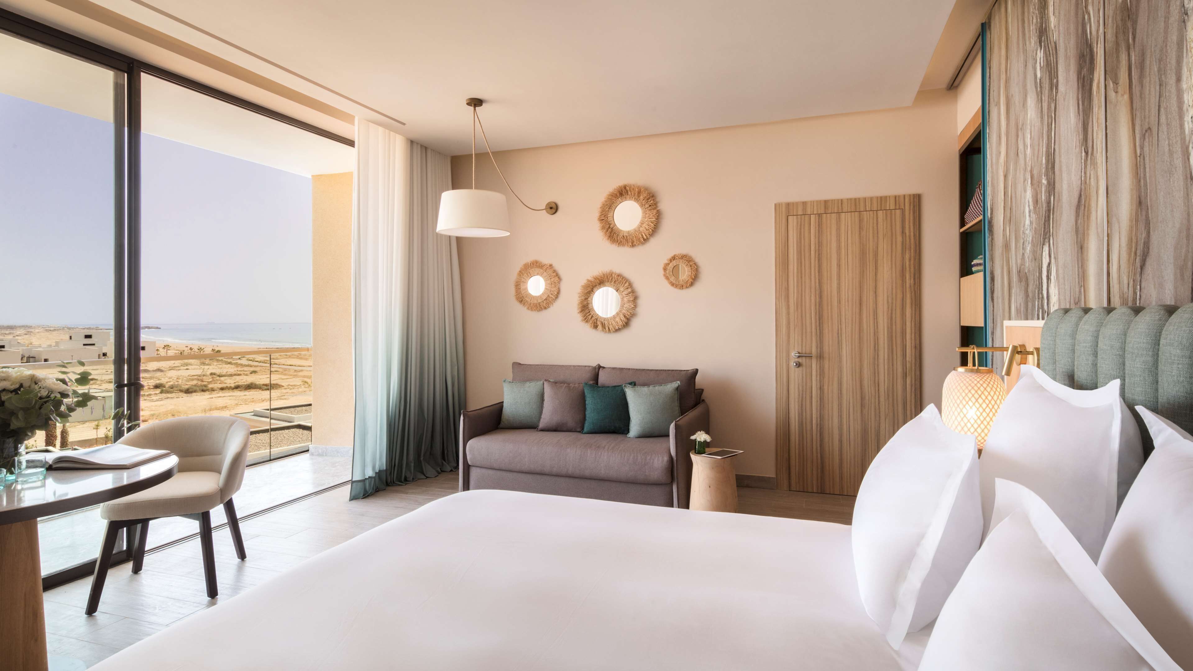 A modern hotel room with a large bed, a sofa, a dining table, and a view of the beach.