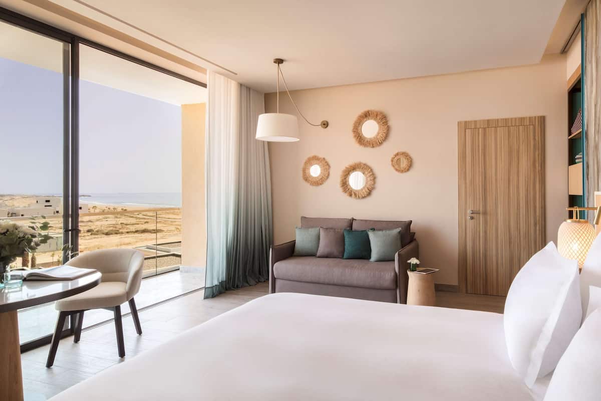 A modern hotel room with a large bed, a sofa, a dining table, and a view of the beach.
