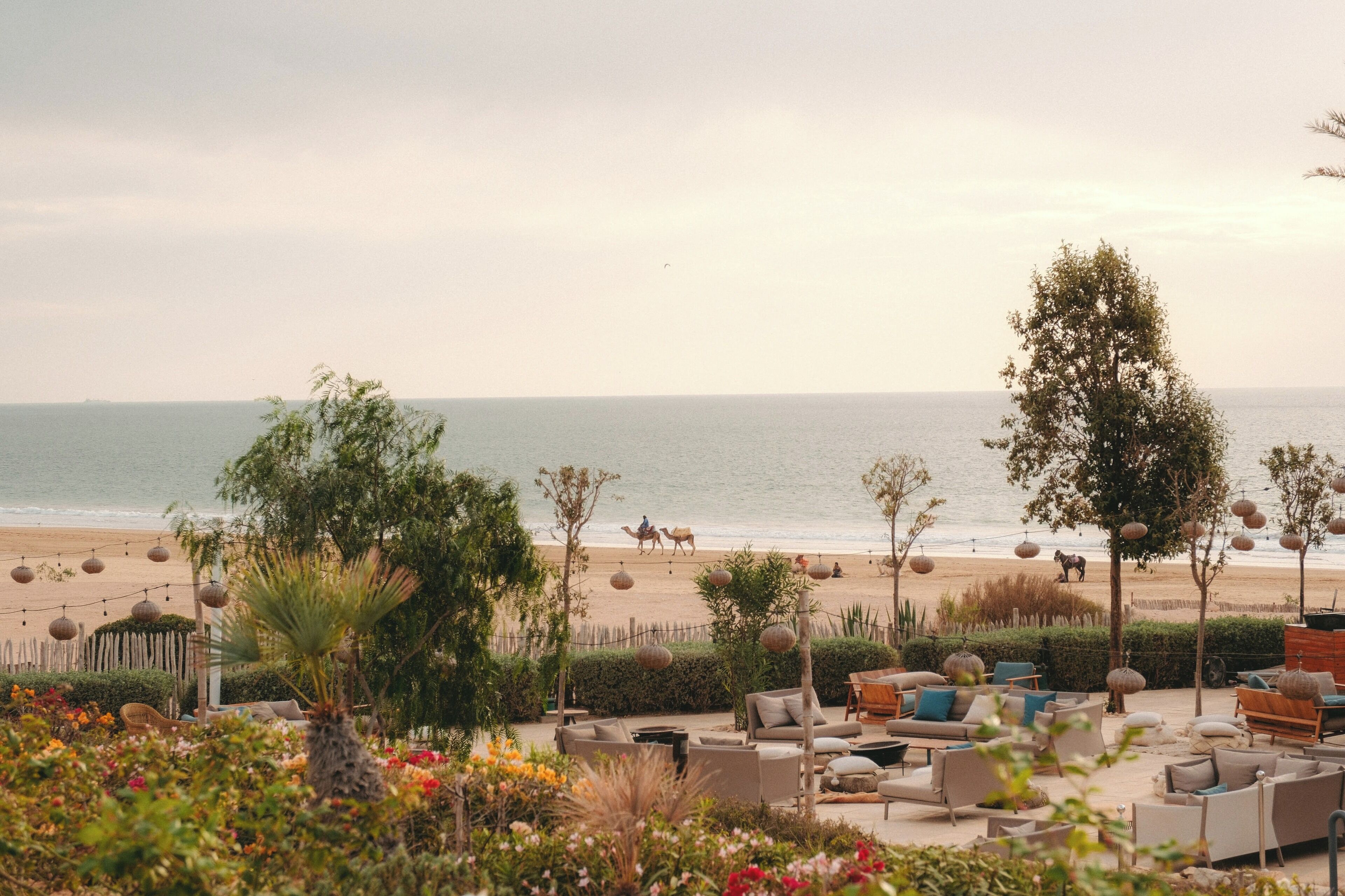 A beachside lounge area with outdoor seating, a view of the sea, and a few people walking on the beach.