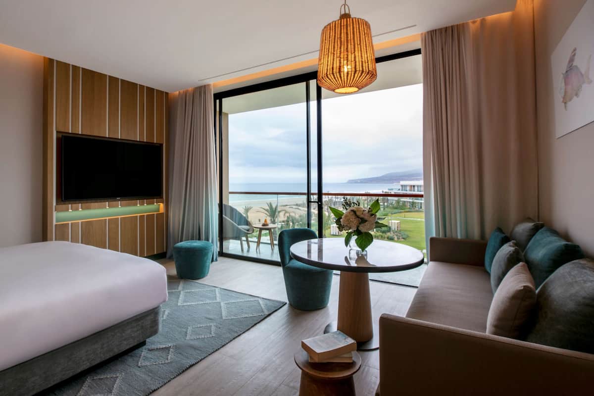 A modern hotel room with a large bed, a sofa, a round table with a vase of flowers, a flat-screen TV mounted on the wall, and a balcony with a view of the sea and buildings.