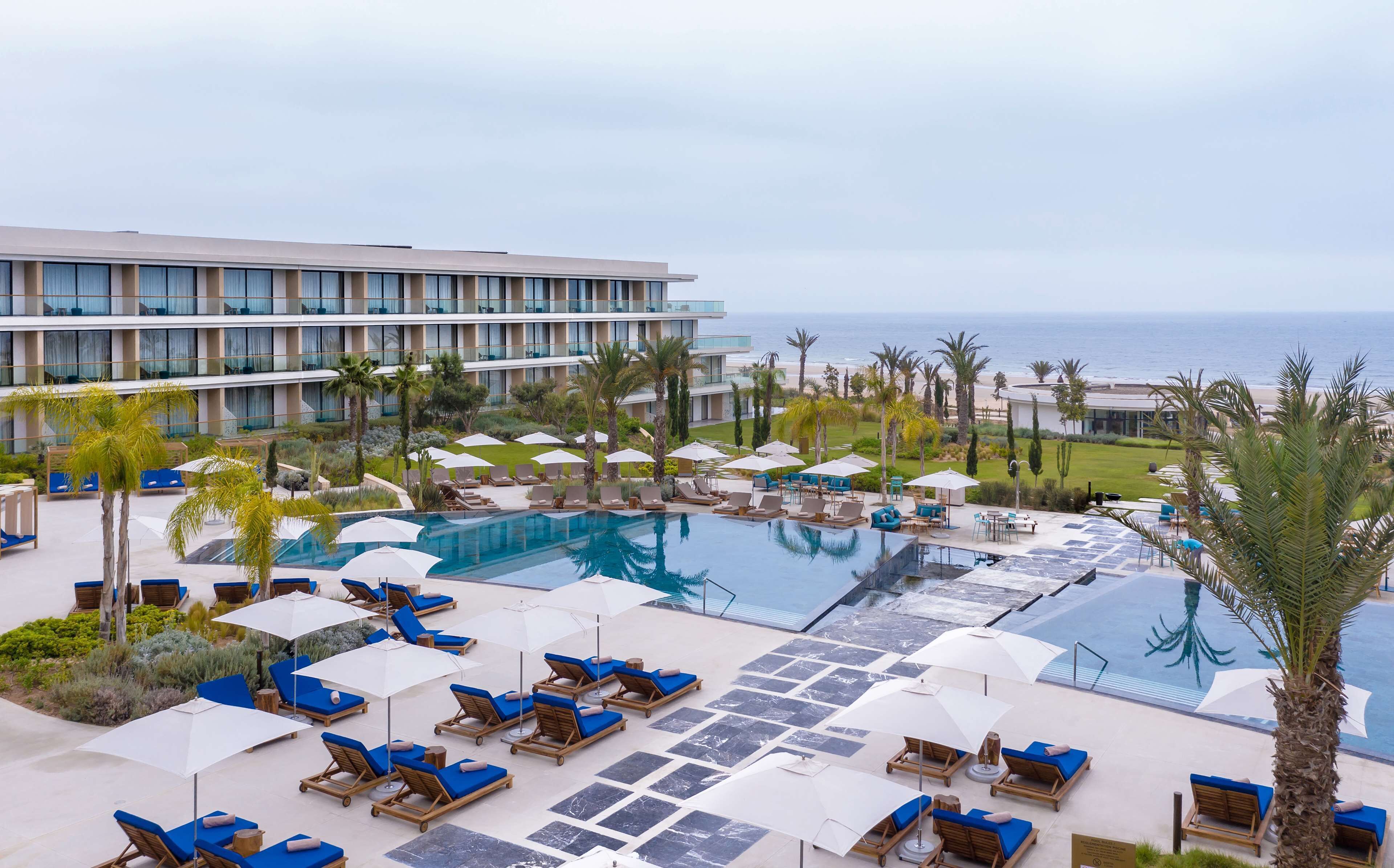 A resort pool area with multiple lounge chairs and umbrellas, a large pool, and a view of the ocean.