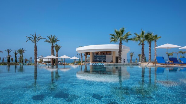 A clear pool with lounge chairs and umbrellas, surrounded by palm trees and a building with a blue and white facade. - Hyatt Regency Taghazout  (Taghazout)