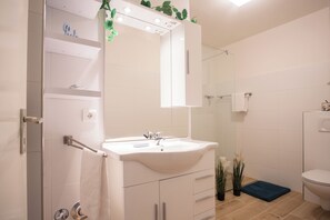 A modern bathroom with a white sink, mirror, and shower. There are plants and a blue mat on the floor.