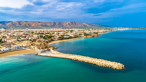 Vista aérea de un pueblo costero con un muelle destacado que se adentra en el mar, rodeado de edificios y un telón de fondo montañoso.