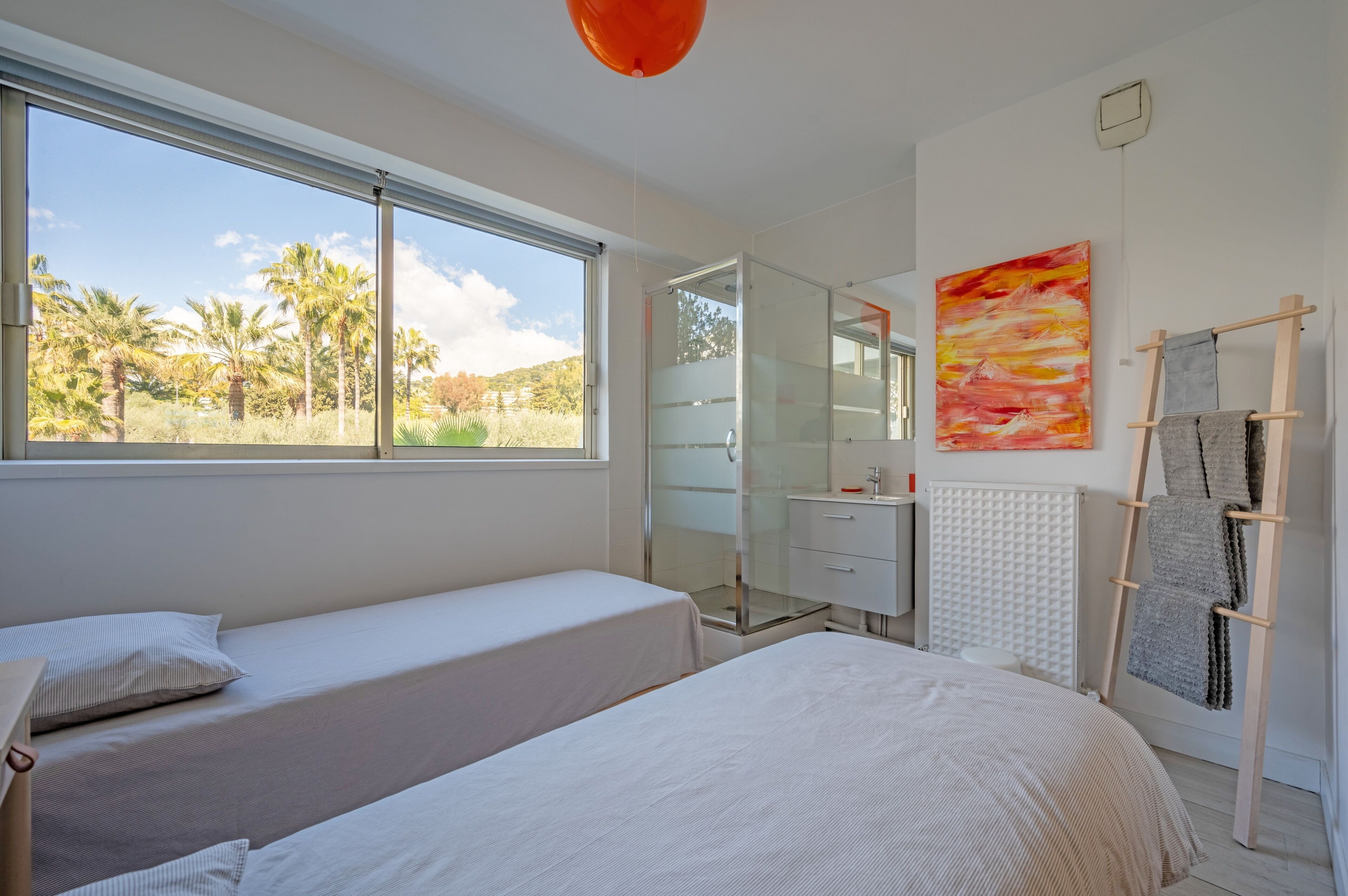Une chambre avec deux lits, une douche en verre, un radiateur et une vue sur des palmiers.