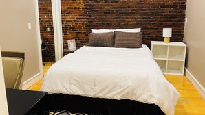 A bedroom with a brick wall, a bed with white bedding, a bedside table with a lamp, and a small shelf. - Boston Harbor views. Modern north end condo.FREE PARKING! (Boston)