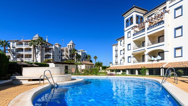 A swimming pool surrounded by multi-story buildings with balconies, palm trees, and a clear blue sky. - EL FARO 60 - One Bedroom Apartment, Sleeps 3 (Punta del moral)