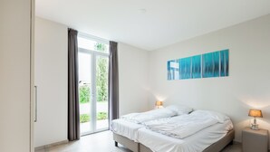 Ein modernes Schlafzimmer mit einem Doppelbett, zwei Nachttischlampen, einem großen Fenster mit Vorhängen und einem blauen abstrakten Gemälde an der Wand.