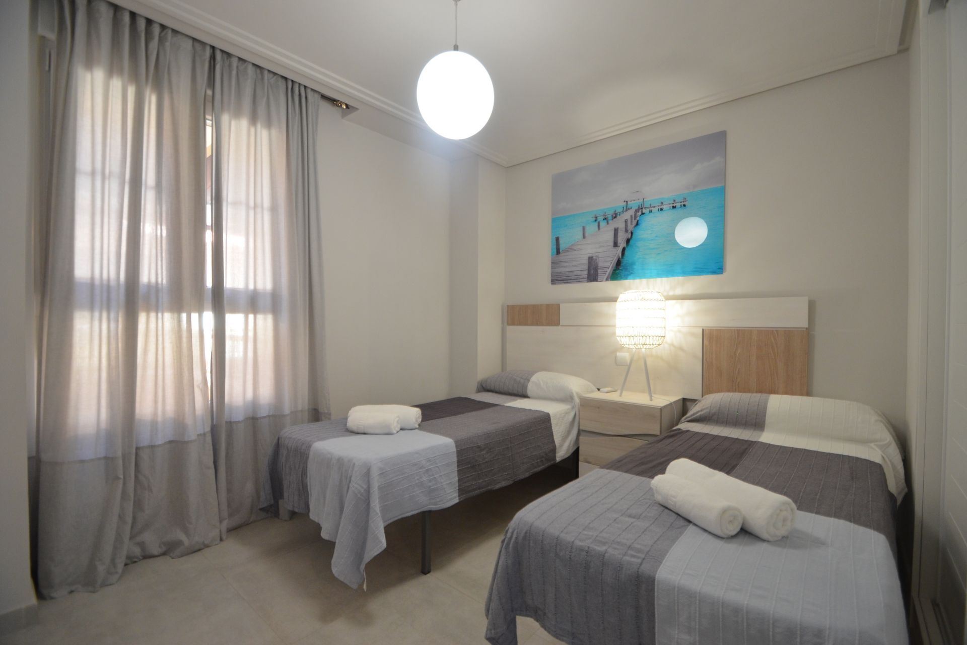 Une chambre d’hôtel avec deux lits, une table de chevet, une lampe, un tableau représentant une jetée et une grande fenêtre avec des rideaux.