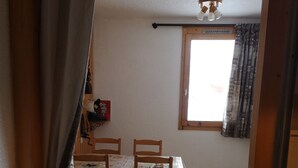 A dining area with a wooden table and chairs, a window with patterned curtains, and a ceiling fan. - Cosy Studio (La Plagne Village)