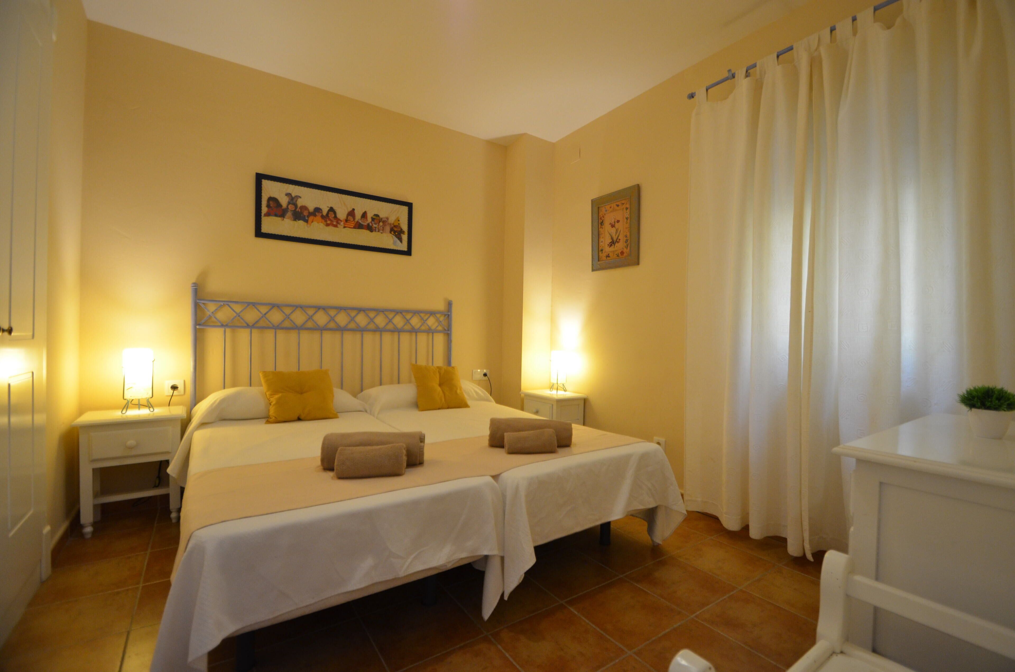 Una cama doble con dos almohadas, una mesita de noche con lámpara, un reloj de pared y una ventana con cortinas.