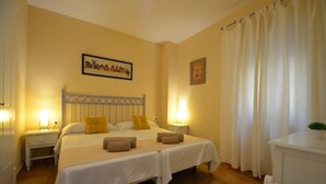 Una cama doble con dos almohadas, una mesita de noche con lámpara, un reloj de pared y una ventana con cortinas.