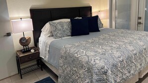 Un lit bien fait, avec une literie à motifs, une lampe de chevet, une petite table de nuit en bois et un coquillage décoratif.