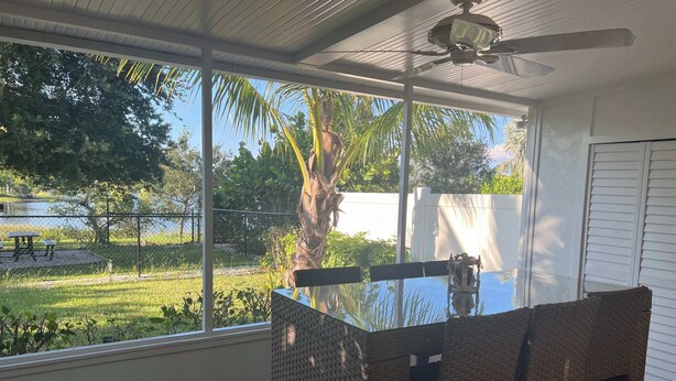 Un coin repas avec une table en verre et des chaises en osier, de grandes fenêtres donnant sur un jardin et un palmier, et un ventilateur de plafond.