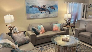 Un salon avec un canapé beige, une table basse en verre et un tableau représentant des tortues de mer accroché au mur.