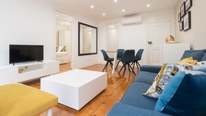 A modern living room with a flat-screen TV, a white coffee table, a blue sofa, and a yellow bench. There is a dining area with a table and chairs.