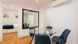 A modern living room with a glass-top dining table, blue upholstered chairs, a large mirror, and a built-in air conditioning unit.