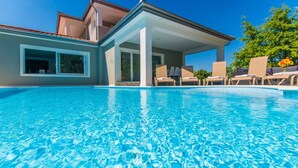 En swimmingpool med krystalklart, blåt vand, omgivet af liggestole og en overdækket terrasse.