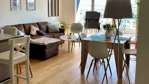 A modern living room with a brown leather sofa, a dining table with chairs, and a large window providing a view of the outdoors. - Appt terrasse vue mer, 2 chambres / Appt terrace sea view with 2 bedrooms (Trouville-sur-Mer)