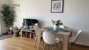 A dining area with a wooden table, white chairs, a potted plant, a framed picture, and a radiator. - Appt terrasse vue mer, 2 chambres / Appt terrace sea view with 2 bedrooms (Trouville-sur-Mer)