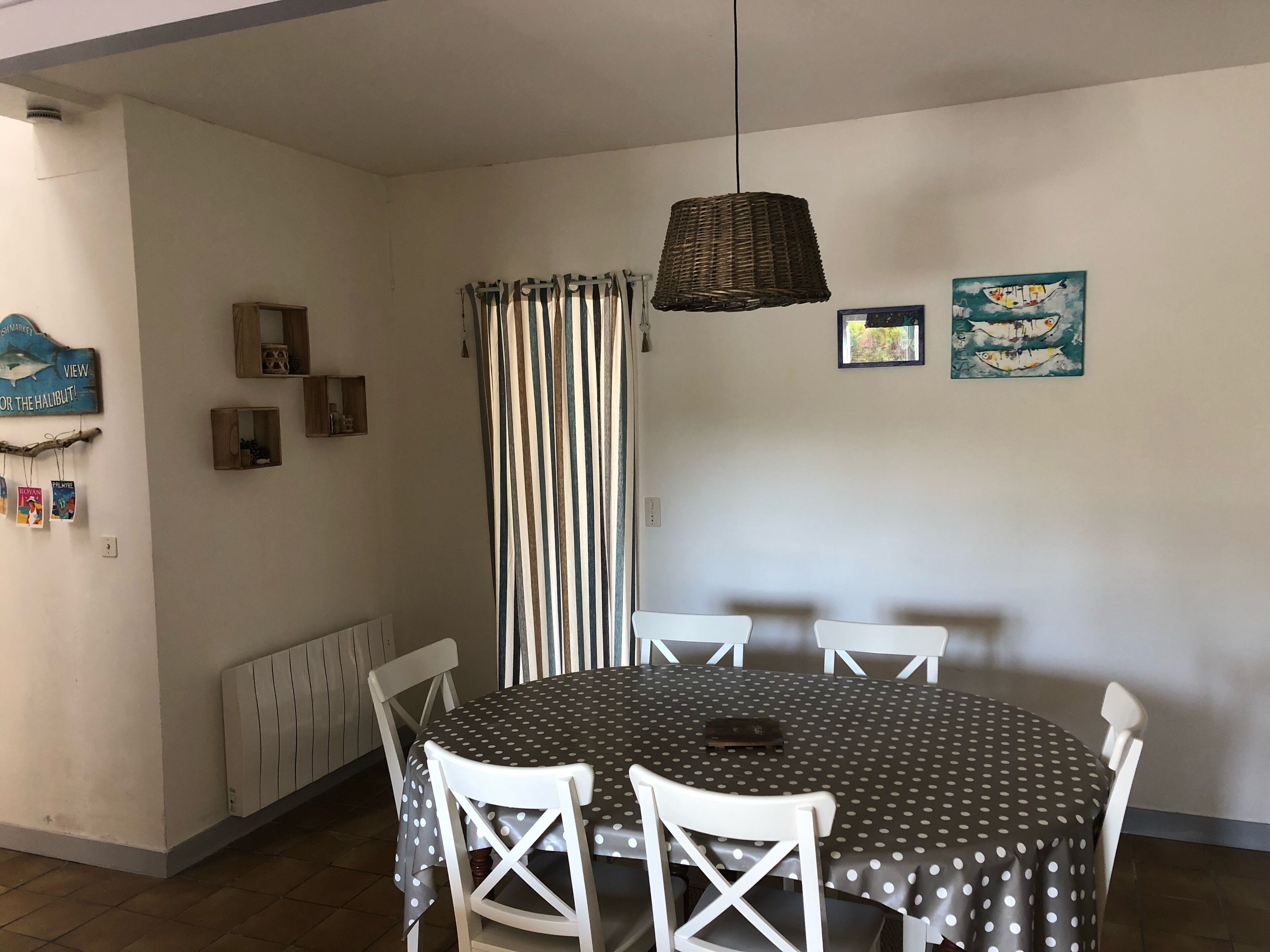 Ein Essbereich mit einem runden Tisch, bedeckt mit einer Tischdecke mit Punkten, umgeben von weißen Stühlen. Über dem Tisch hängt eine Körbeschranke als Lichtquelle.