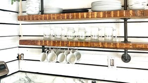 Una cocina con paredes de madera blanca, encimera de mármol y estantes con vajilla, tazas y una planta.