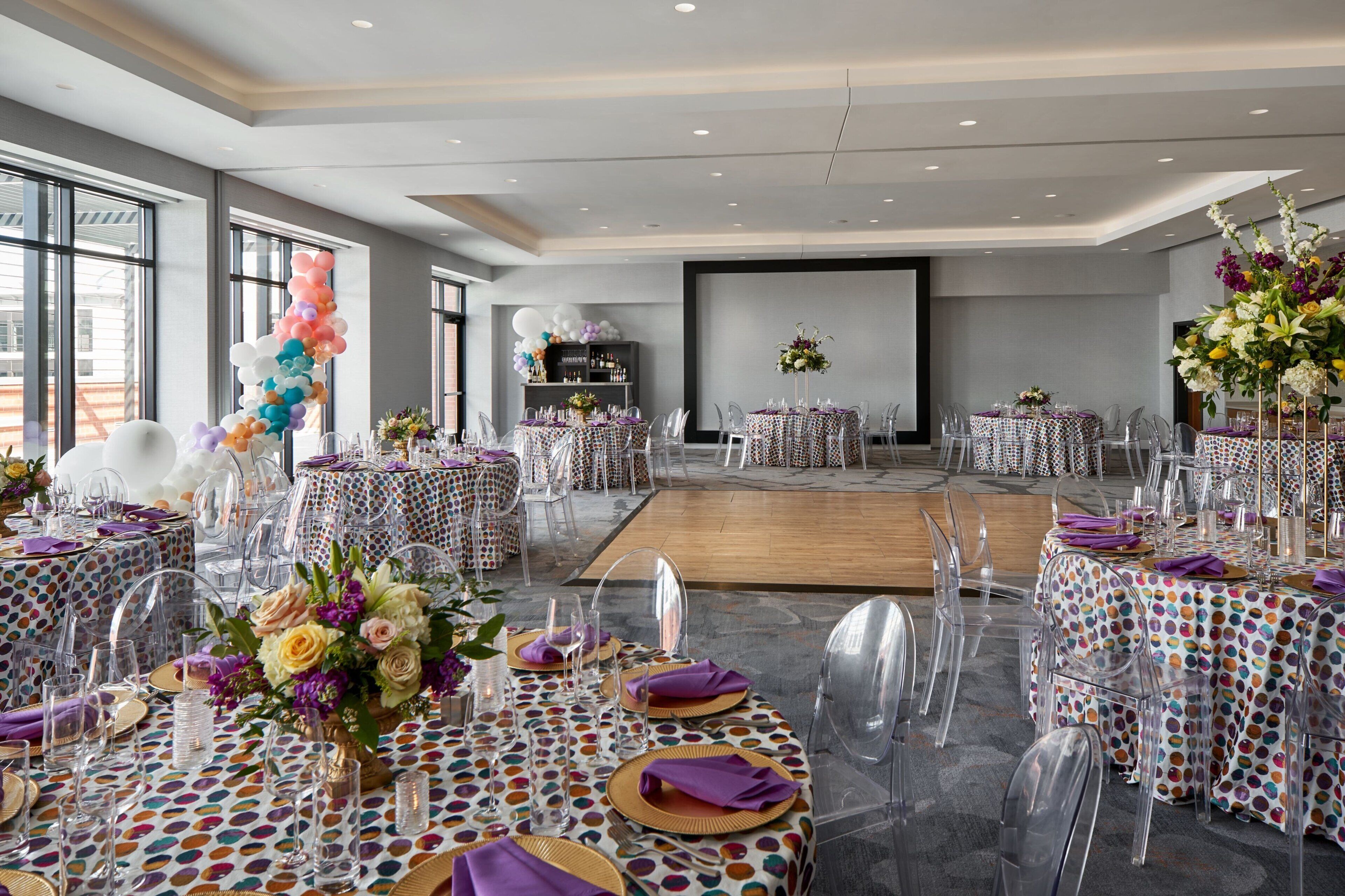 Un amplio salón de banquetes con mesas redondas, sillas transparentes y diversos centros de mesa florales.