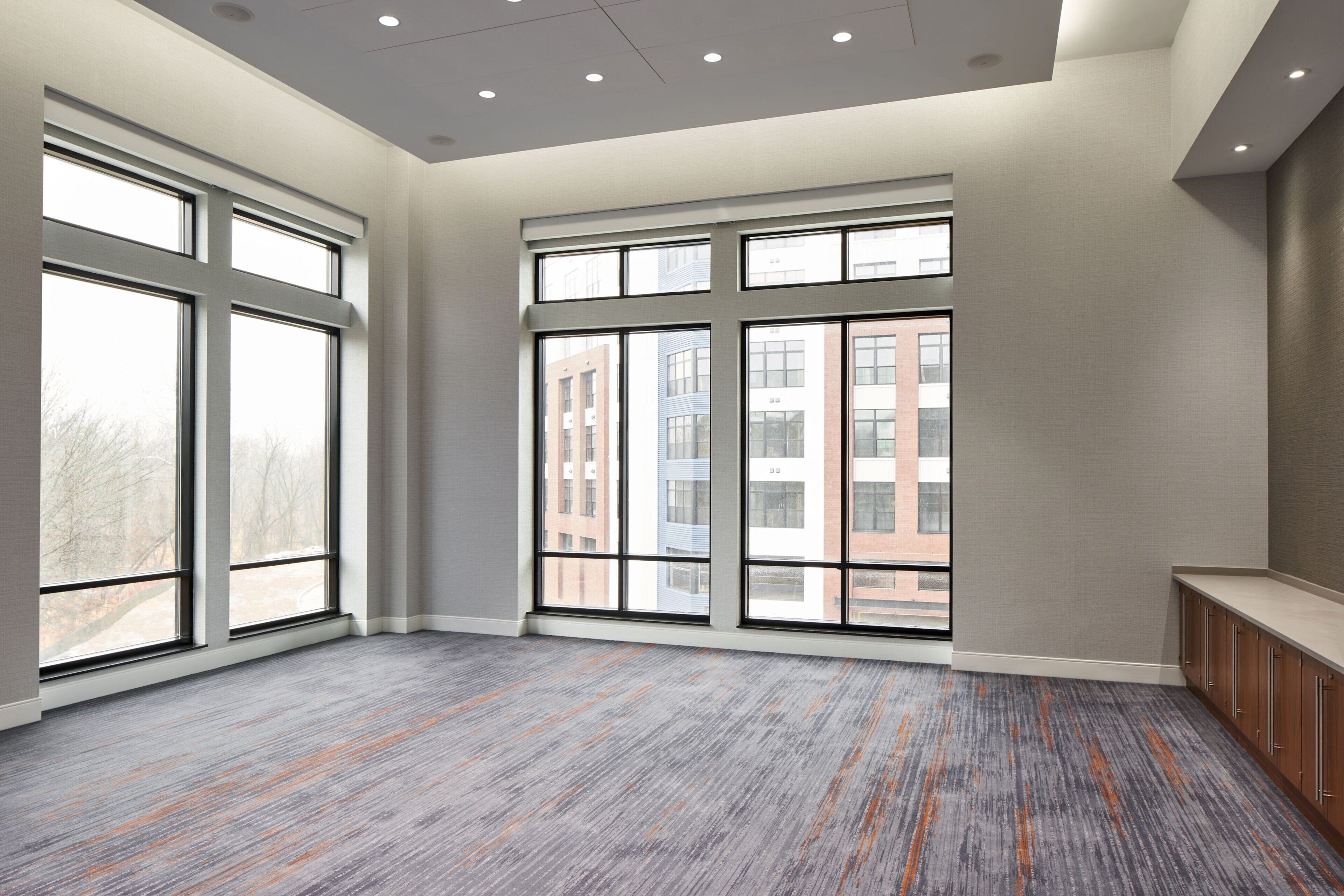 A spacious room with large windows, a grey carpet, and a wooden bench along the wall.