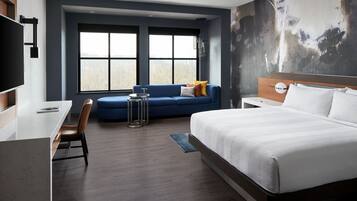 Una habitación de hotel moderna con una cama grande, un escritorio, un sofá azul y vistas al exterior a través de amplios ventanales.