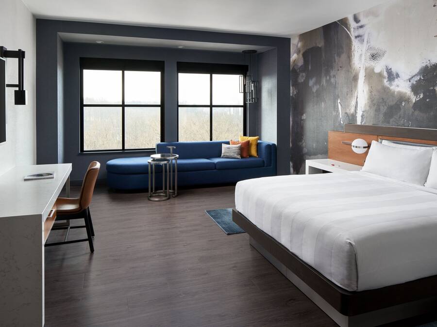 Una habitación de hotel moderna con una cama grande, un escritorio, un sofá azul y vistas al exterior a través de amplios ventanales.