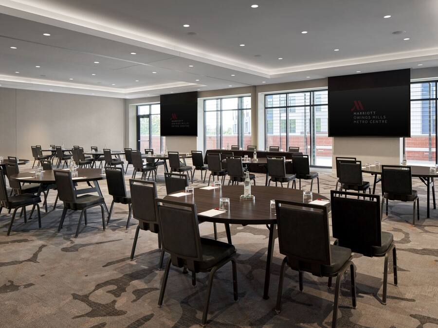 Sala de conferência com várias mesas e cadeiras, janelas amplas e telas exibindo o logotipo Marriott.