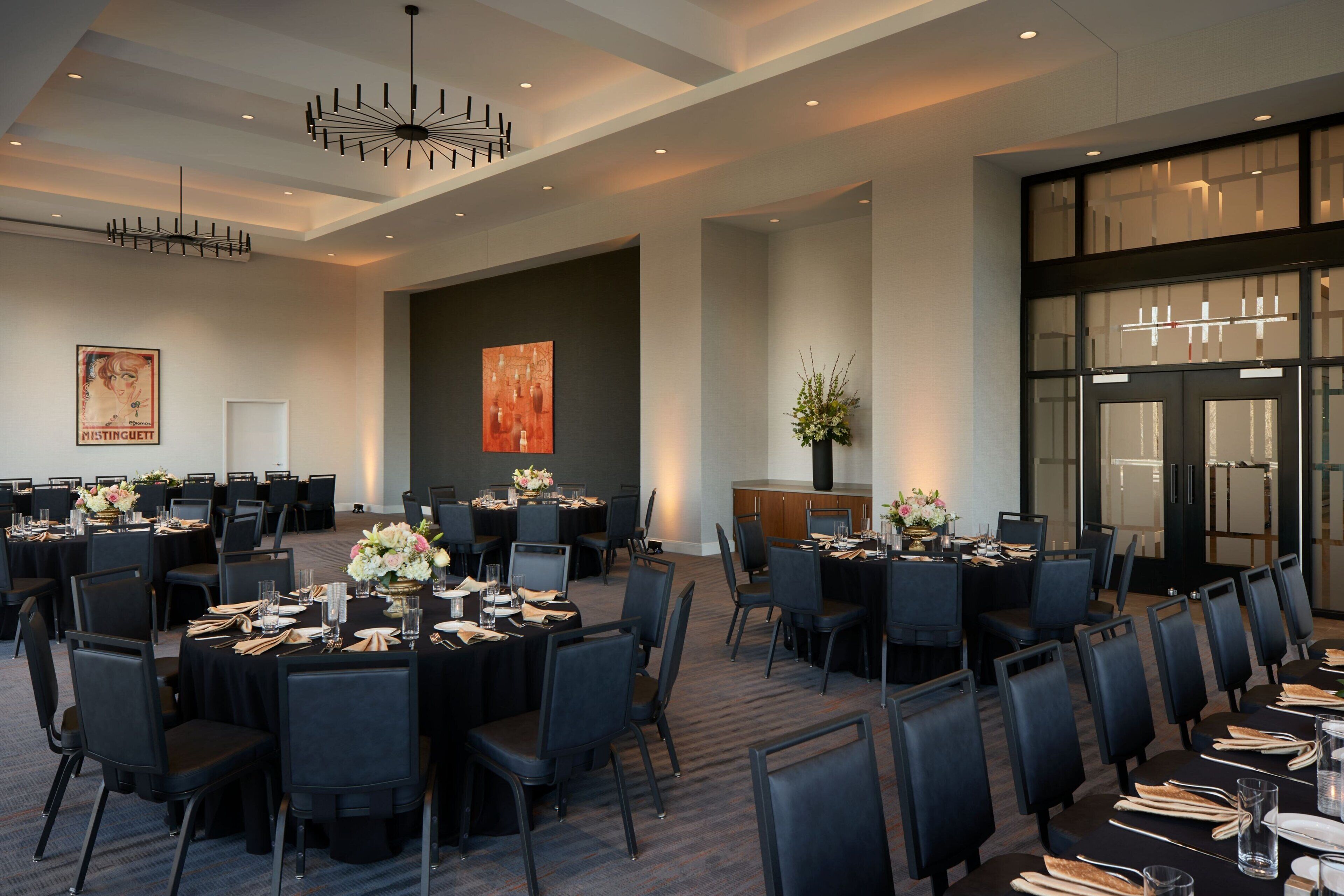 A large conference room with multiple tables set for dining, featuring black chairs and tables, a chandelier, and a large window with blinds.
